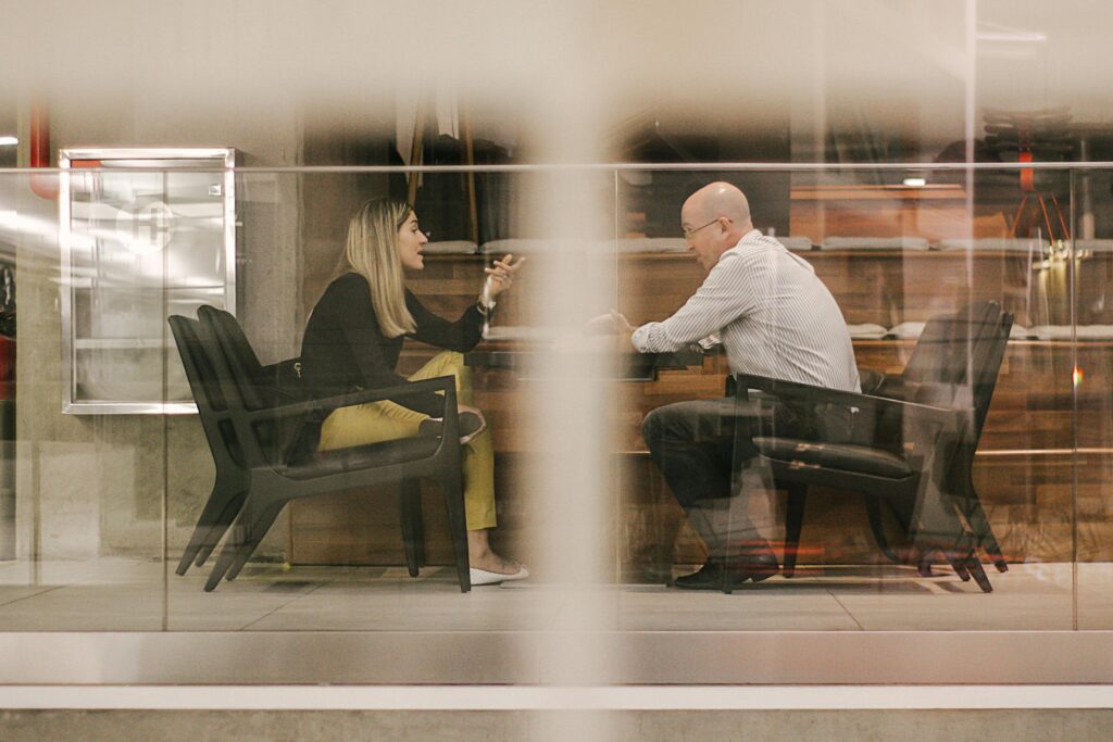 Two professionals engaged in a discussion in a modern office setting through a glass window.